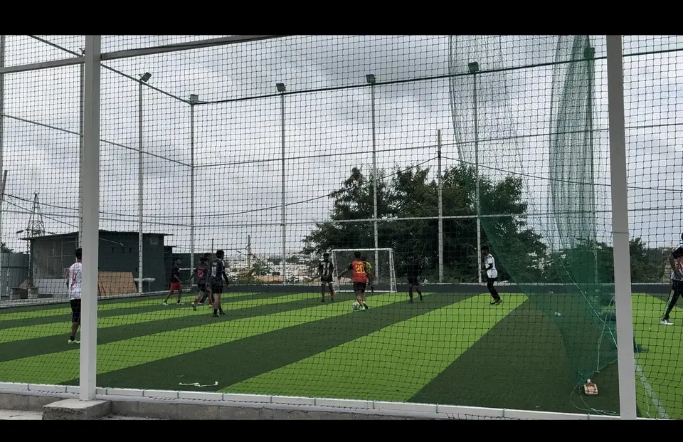 Sports Aura in daylight seen through the net with white poles, floodlights, striped turf and distant trees.